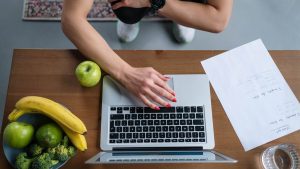Overhead view of hands on laptop with fruits, vegetables, and paper on wooden table.